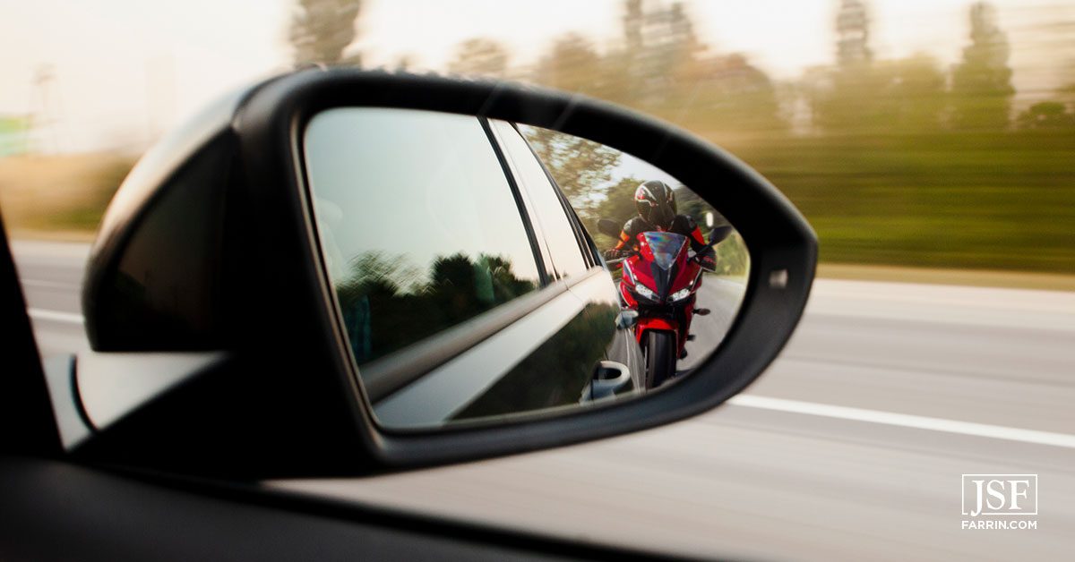 Motorcycle driver reflection in the side-view mirror of a car driving on a Durham road.