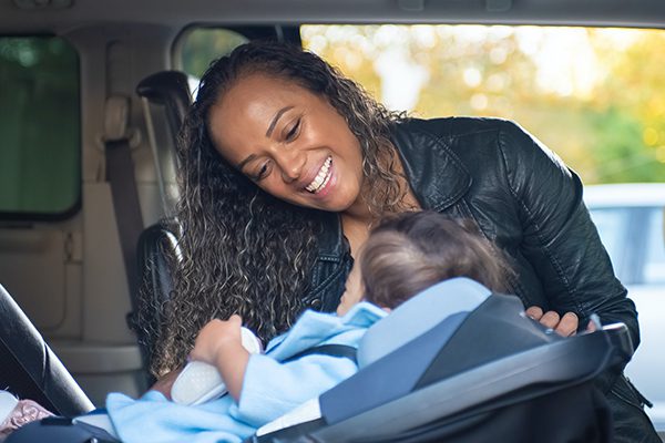 A smiling black mother buckles her baby into his carseat.