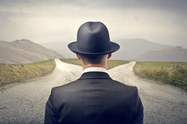 Conceptual image of a man in a black suit and hat looking at a fork in the road.