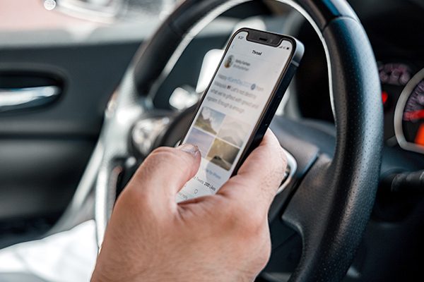 A hand holding a cell phone in front of a steering wheel while driving a car.