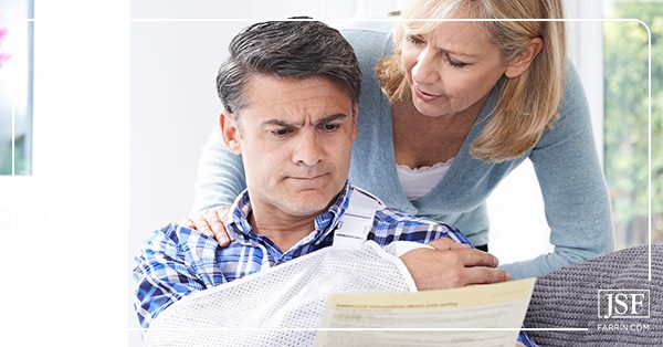Man with arm in sling frowns at denial letter while wife consoles him with hands on his shoulders